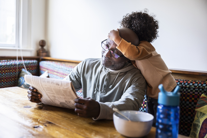 Father reading newspaper while toddler climbs on him- poverty 